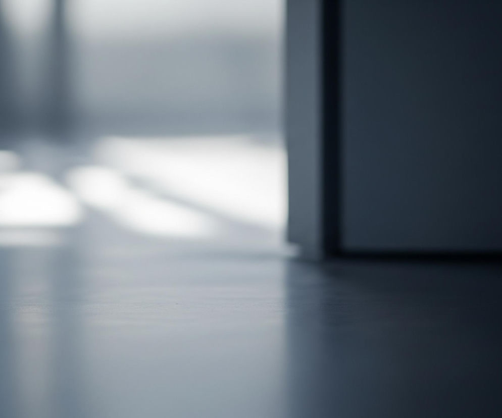 Podłoga w jasnoszarym odcieniu rozciąga się w kierunku centrum kadru, delikatnie jaśniejąc w jego lewej, środkowej części. Z prawej strony kadru widoczna jest ciemnoszara ściana lub futryna drzwi, tworząca pionowy, rozmyty element. W tle, niewyraźne, jasne kształty sugerują źródła światła lub odbicia na dalszej powierzchni. Całość obrazu utrzymana jest w chłodnej tonacji szarości i bieli, z efektem bokeh, co sprawia, że jedynie pierwszy plan jest ostry, a reszta rozmyta.