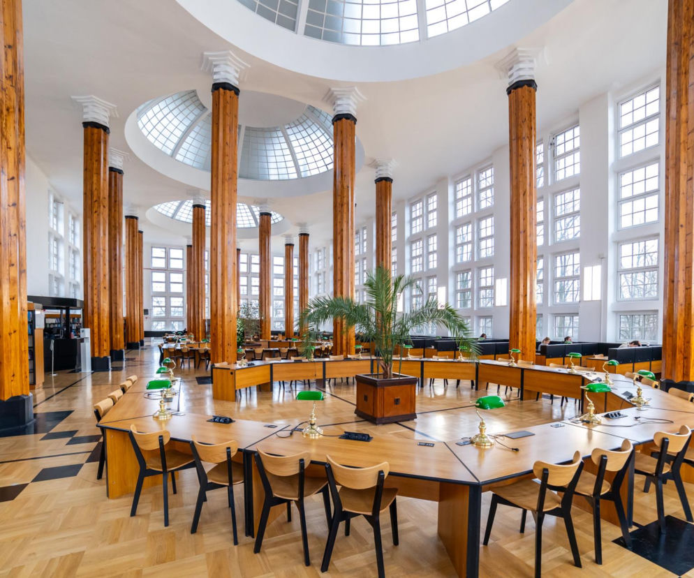 Wnętrze modernistycznej Czytelni Głównej Biblioteki SGH w Warszawie, projektu Jana Koszczyca-Witkiewicza, z charakterystycznymi drewnianymi kolumnami, licznymi oknami, świetlikami i oryginalnymi stołami do czytania. O architekturze tego wyjątkowego gmachu przeczytasz na Architektura Murator Plus.