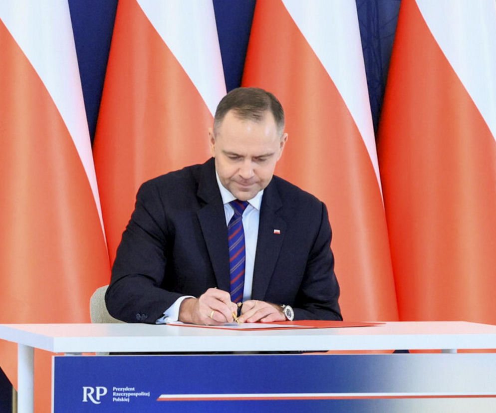 Karol Nawrocki, prezydent RP, podpisuje dokumenty, siedząc przy białym biurku z herbem Polski. W tle widoczne są flagi Polski. O szczegółach ustawy o Państwowej Inspekcji Pracy, która wywołuje wiele emocji, przeczytasz na Super Biznes.