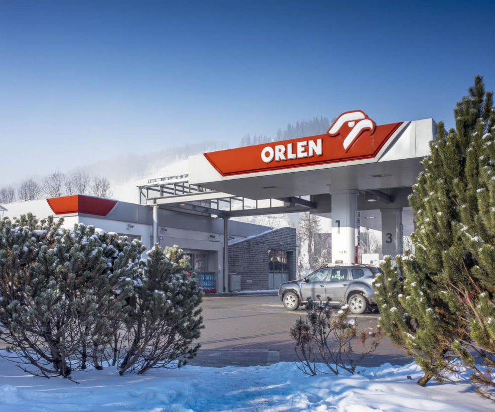 Stacja paliw ORLEN w zimowej scenerii, z widocznym dachem i logo firmy, a także samochodem na podjeździe. W tle ośnieżone drzewa i góry pod błękitnym niebem. O promocji na paliwo ORLEN przeczytasz na portalu Super Biznes.