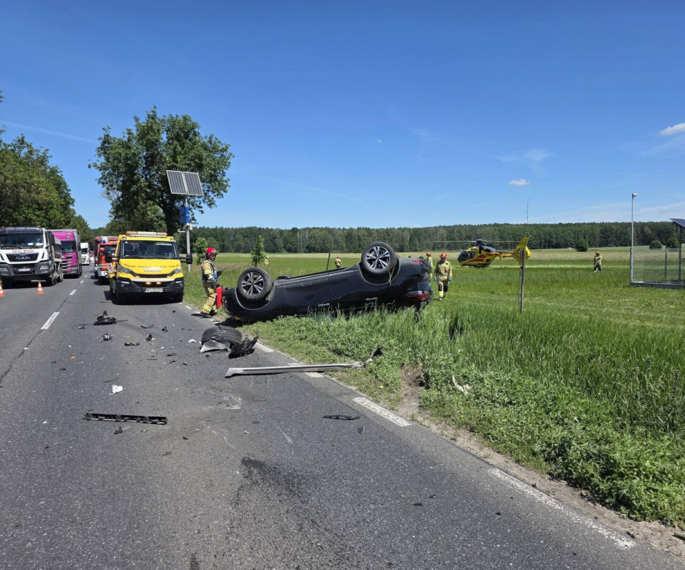 Czarny samochód osobowy leży przewrócony na dach na poboczu drogi krajowej nr 63 w Chodowie. Na miejscu wypadku pracują służby ratunkowe, w tym strażacy i LPR. Szczątki pojazdów leżą rozrzucone na asfalcie. Więcej o zdarzeniu na naszym portalu.