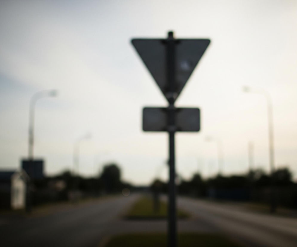 Centralnie, na pierwszym planie, znajduje się rozmyty znak drogowy składający się z ciemnej, odwróconej trójkątnej tablicy i mniejszej, prostokątnej pod nią, osadzonych na słupie. Tło stanowi szeroka droga biegnąca w perspektywie, z pasem zieleni pośrodku. Po obu stronach drogi widać rozmazane kształty budynków, latarni ulicznych i drzew, a horyzont rozświetla jasne, rozmyte słońce, tworząc silną poświatę po prawej stronie.
