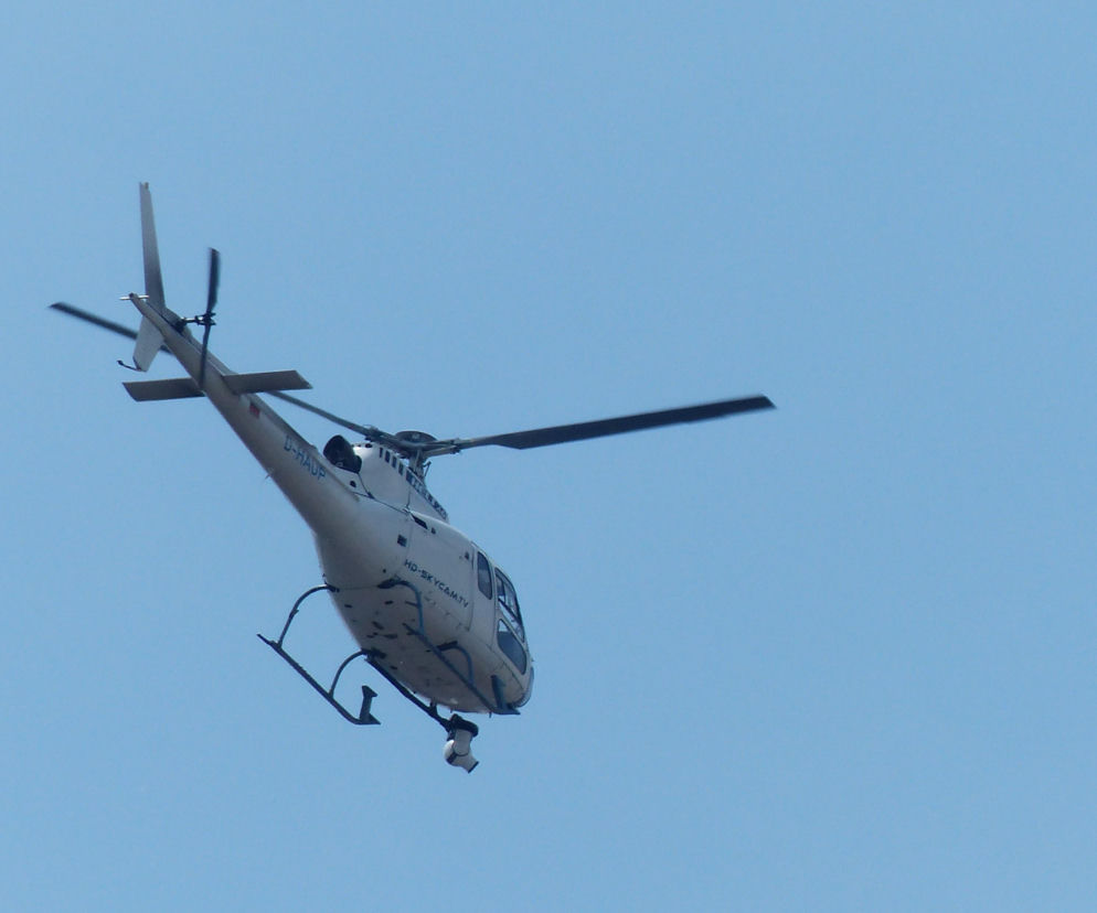 Biały helikopter z czarnymi łopatami wirnika i podwoziem, z zamontowaną kamerą pod kadłubem, przelatuje na tle czystego, błękitnego nieba. Maszyna wspiera obławę policyjną pod Wadowicami, a o szczegółach akcji można przeczytać na naszym portalu.