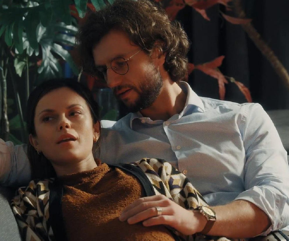 Paulina Lasota (Kasia) i Mateusz Mosiewicz (Mariusz) na kanapie, on obejmuje ją, patrząc z czułością na jej ciążowy brzuszek. Kobieta ma na sobie brązowy sweter i wzorzystą narzutkę, on jasnoniebieską koszulę. Para z M jak miłość przeżywa kryzys, o którym można przeczytać na Super Seriale.