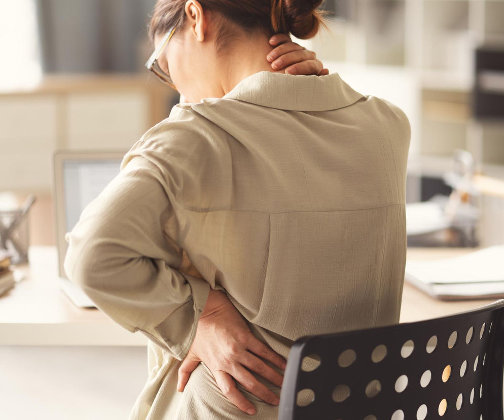Kobieta z bólem pleców i szyi, masująca się po pracy przy biurku. Długotrwała praca zdalna bez ergonomicznego stanowiska często prowadzi do syndromu kanapowego biura. Na Poradnik Zdrowie znajdziesz sposoby na walkę z bólem.