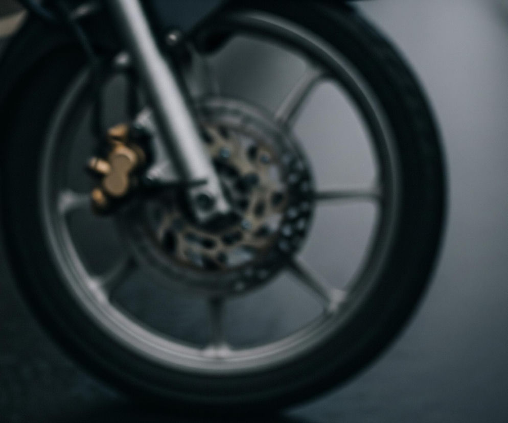 Przednie koło motocykla znajduje się na pierwszym planie, z dominującą oponą i srebrną felgą szprychową. W centralnej części felgi widoczna jest złota tarcza hamulcowa z widocznymi perforacjami i jasnym, mosiężnym zaciskiem. Górna część zdjęcia jest rozmyta, ukazując elementy zawieszenia oraz ciemne tło. Kolory są głównie ciemne i stonowane, od głębokiej czerni po różne odcienie szarości i metalicznego srebra.