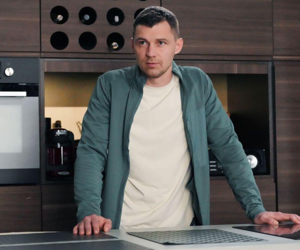 Mężczyzna w zielonej bluzie i jasnym T-shircie, Darek (Andrzej Niemyt), stoi w kuchni, opierając się o blat, z zamyślonym wyrazem twarzy. Przygotowuje się do wyjazdu do Lizbony w serialu Barwy szczęścia, co będzie przełomową decyzją po śmierci Karola. Więcej o losach bohaterów na Super Seriale.