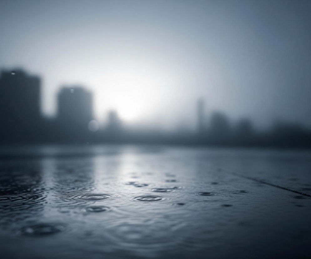Krople deszczu uderzają o ciemną, wilgotną powierzchnię na pierwszym planie, tworząc okręgi fal na wodzie. W tle widoczna jest rozmyta panorama miasta, z kilkoma wysokimi budynkami o niewyraźnych konturach, tonąca w mglistej atmosferze. Jasne światło przebija się przez chmury w centralnej części horyzontu, rzucając delikatny blask na miejski krajobraz. Całość utrzymana jest w chłodnej tonacji szarości i błękitu.