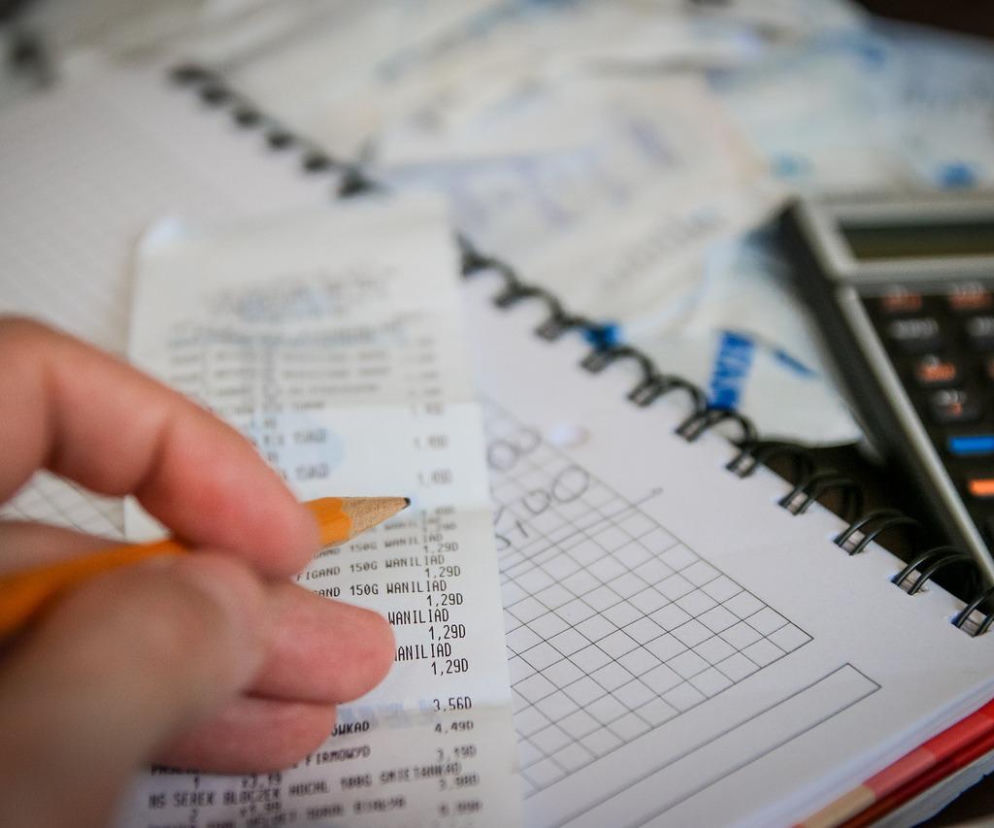 Ręka z ołówkiem analizująca długi paragon, obok notatnik w kratkę i kalkulator. Symbolizuje wysokie rachunki za prąd i ogrzewanie, co skłania rolników do szukania oszczędności, o czym przeczytasz na Super Biznes.