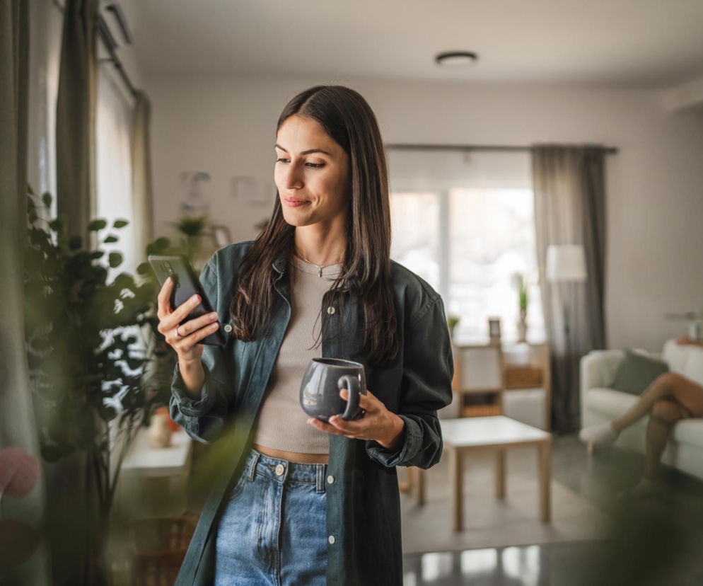 Młoda kobieta z długimi, ciemnymi włosami, w dżinsach i ciemnozielonej koszuli, trzymająca smartfon i kubek, stoi przy oknie. W tle widoczny mężczyzna na sofie. Zdjęcie ilustruje temat smartphone pinky i jak zapobiegać bólowi dłoni, o czym przeczytasz na Poradniku Zdrowie.