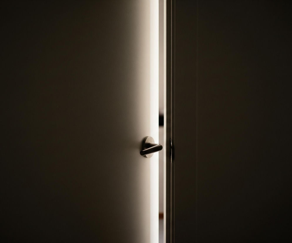Ciemne drzwi uchylone są nieznacznie, ukazując jasne, pionowe pasmo światła pośrodku kadru. Po lewej stronie, na ciemnej powierzchni drzwi, widoczna jest okrągła, metalowa klamka w kolorze grafitowym, zwrócona lekko w dół. Po obu stronach pasa światła rozciągają się ciemne, niemal czarne powierzchnie ścian lub skrzydeł drzwi, z delikatnymi, widocznymi teksturami.