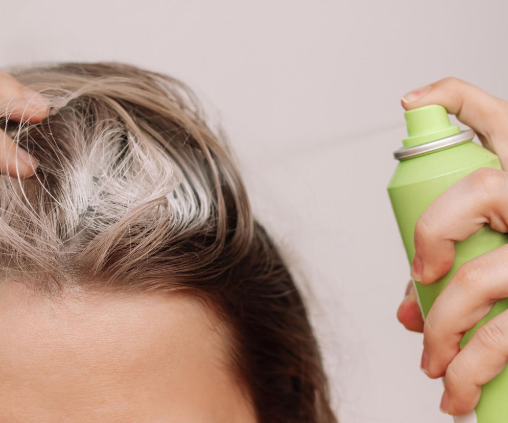 Kobieta w akcji użycia suchego szamponu, rozpylając go na odrosty swoich włosów, co ilustruje temat błędów w stosowaniu tego kosmetyku, które mogą prowadzić do podrażnień i łupieżu. Więcej na temat bezpiecznego używania suchego szamponu przeczytasz na Poradnik Zdrowie.