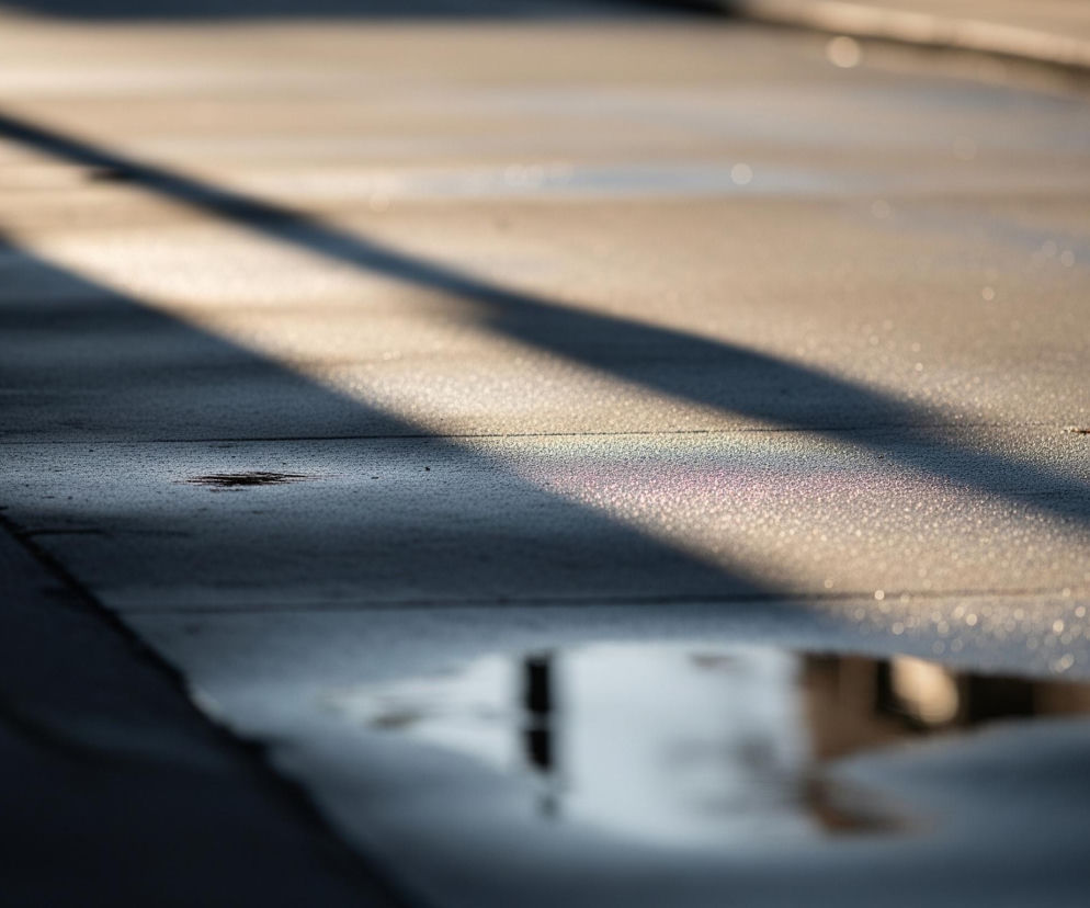 Na betonowej powierzchni drogi rozciągają się ukośne cienie, tworząc kontrastujące pasy ciemnego i jasnego koloru. Na pierwszym planie, w prawym dolnym rogu, widoczna jest kałuża wody odbijająca rozmyty obraz jasnych, niewyraźnych kształtów. Fragment powierzchni w lewej górnej części zdjęcia jest jaśniejszy i pokryty drobnymi, błyszczącymi kropelkami, a w jej środkowej części, w obrębie cienia, dostrzegalne są delikatne, kolorowe smugi, przypominające tęczę.