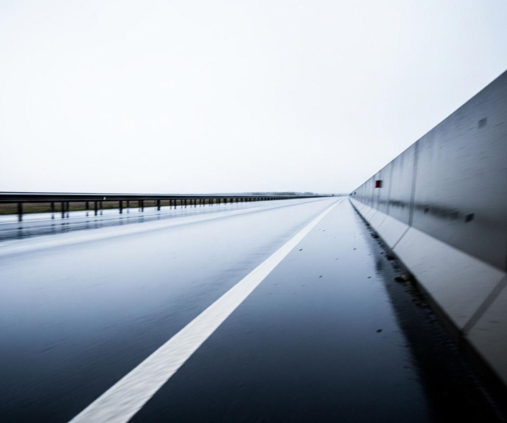 Asfaltowa droga, mokra od deszczu, rozciąga się w głąb obrazu, zajmując większą część dolnej i środkowej kompozycji. Na lewo od niej widać metalową barierę ochronną, a po prawej pionową, szarą ścianę z widocznymi czerwonymi elementami odblaskowymi. Niebo jest jednolicie jasne, biało-szare, bez widocznych chmur, co nadaje zdjęciu ogólny chłodny odcień.