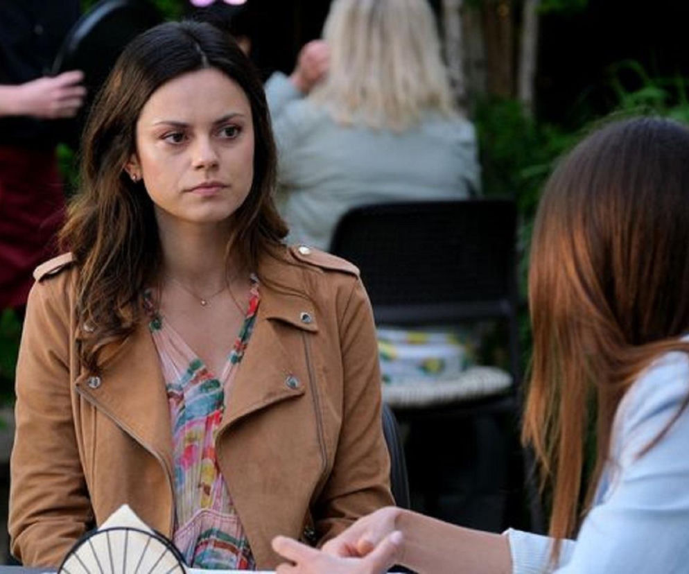 Kasia (Paulina Lasota) siedzi przy stoliku, patrząc z zaniepokojeniem w bok, ma na sobie brązową kurtkę i wzorzystą bluzkę. Przed nią widoczne są dłonie drugiej kobiety, co odzwierciedla napiętą sytuację z 1896 odcinka M jak miłość. Więcej o tym na Super Seriale.