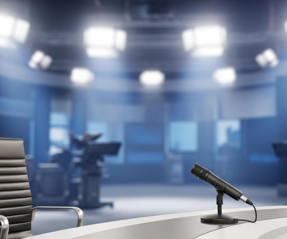 W centrum obrazu znajduje się puste, nowoczesne studio telewizyjne z białym, zakrzywionym biurkiem na pierwszym planie. Na blacie biurka stoi czarny mikrofon na podstawce, podłączony do kabla. Po lewej stronie biurka widoczne jest czarne, skórzane krzesło biurowe z metalowymi podłokietnikami i ramą, ustawione tyłem do widza. W tle, rozmyte, widoczne są niebieskie ściany, ekrany i ciemne sylwetki dwóch kamer studyjnych na statywach, a sufit oświetlają jaskrawe, prostokątne reflektory.