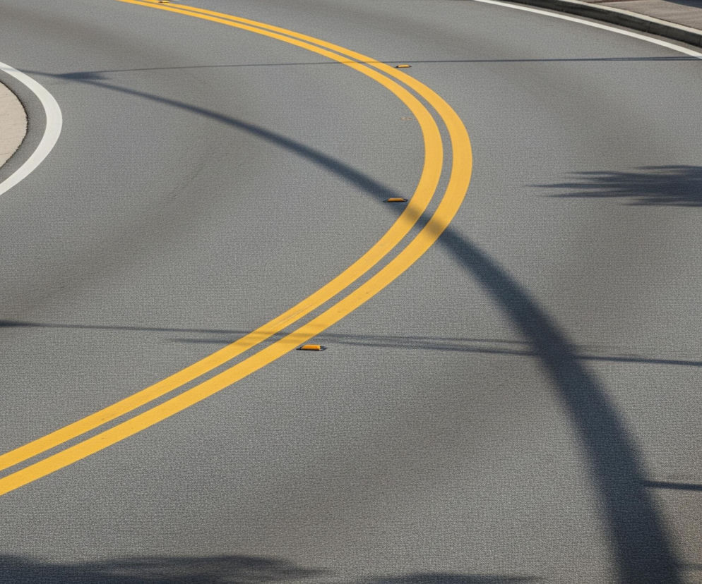 Szara asfaltowa droga zakręca w prawo, z podwójną, ciągłą żółtą linią wyznaczającą jej środek. Wzdłuż lewej krawędzi jezdni biegnie biała linia, oddzielająca ją od betonowego krawężnika i chodnika z kratkami ściekowymi. Na drodze widoczne są cztery żółte odblaski, a także długie, ciemne cienie, prawdopodobnie pochodzące od słupów lub drzew, przecinające asfalt.