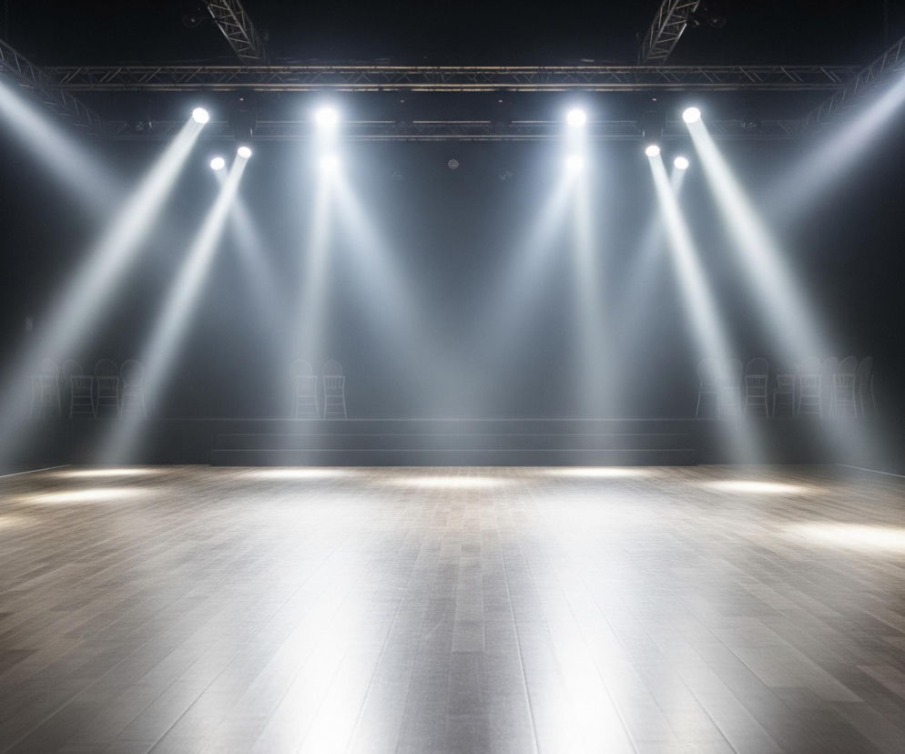 Centralna część obrazu przedstawia oświetloną scenę z drewnianą podłogą, na której widoczne są jasne, owalne plamy światła. Z góry, na czarnym tle, rozmieszczone są reflektory emitujące intensywne, białe snopy światła, które przecinają lekko zamglone powietrze. W tle, na podwyższeniu, znajdują się rzędy krzeseł, a po bokach sceny również widać krzesła ustawione wzdłuż ciemnych ścian.