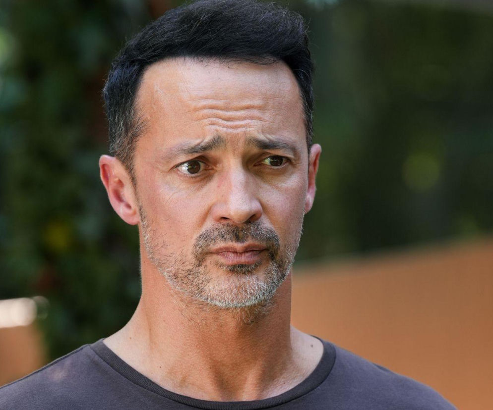 Zbliżenie na Mariusza (Rafał Cieszyński) z serialu Barwy szczęścia z zmartwionym wyrazem twarzy, szare tło z zielonymi liśćmi. Mariusz w 3305 odcinku serialu pozna prawdę o intrydze Janki. Więcej o losach bohatera przeczytasz na stronie Super Seriale.