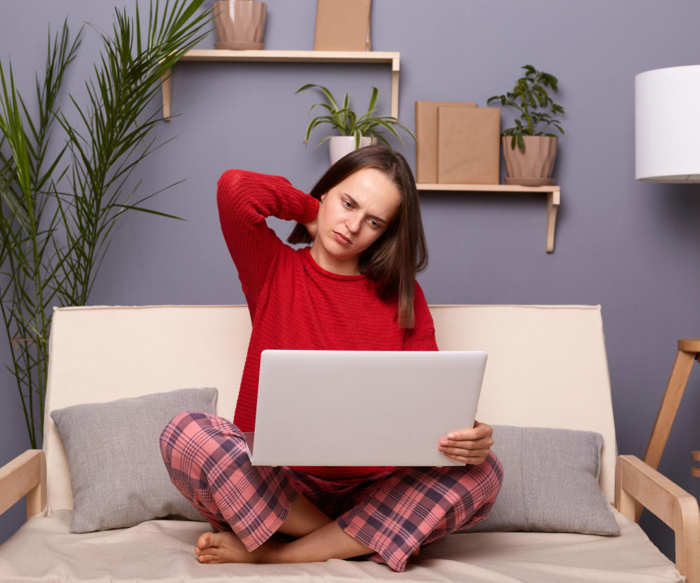 Kobieta w czerwonym swetrze i piżamowych spodniach, siedząca na kanapie z laptopem, masuje kark z wyrazem dyskomfortu, ilustrując problem syndromu tekstowej szyi. Na Poradnik Zdrowie można przeczytać, jak praca w niewłaściwej pozycji obciąża kręgosłup.