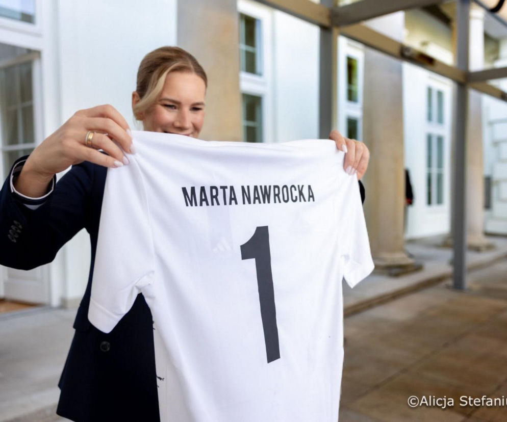 Uśmiechnięta Marta Nawrocka w eleganckiej marynarce trzyma białą koszulkę piłkarską z nazwiskiem Marta Nawrocka i numerem 1. Scena nawiązuje do spotkania z młodymi piłkarzami, o którym można przeczytać na naszym portalu.