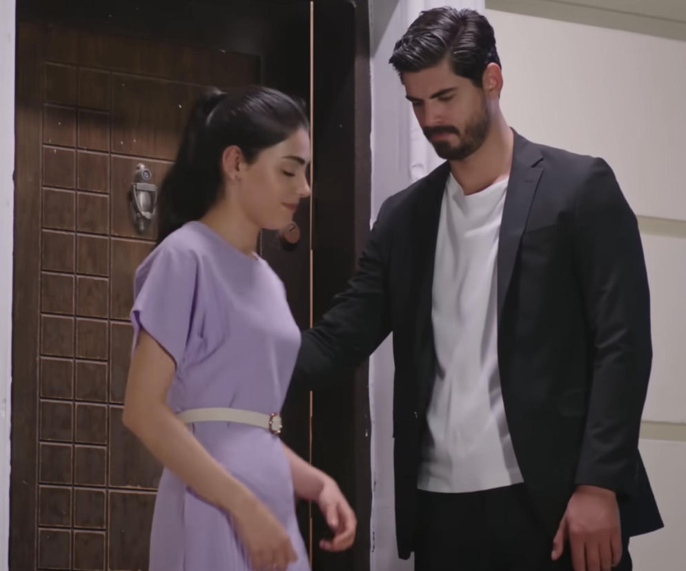 Hancer, ubrana w liliową sukienkę z białym paskiem, i Cihan w czarnej marynarce z białą koszulką, stojący w progu drzwi, w emocjonalnej scenie powrotu do domu w tureckiej telenoweli „Panna Młoda”, o której możesz przeczytać na portalu Super Seriale.