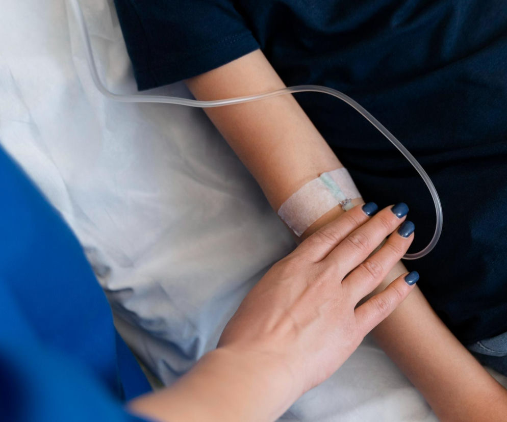 Ręka pacjenta z cewnikiem dożylnym, który jest przytrzymywany przez dłoń w granatowych paznokciach. Na ramieniu widoczny opatrunek i dren, symbolizujące dostęp naczyniowy do dializ, o którym więcej przeczytasz na Poradnik Zdrowie.
