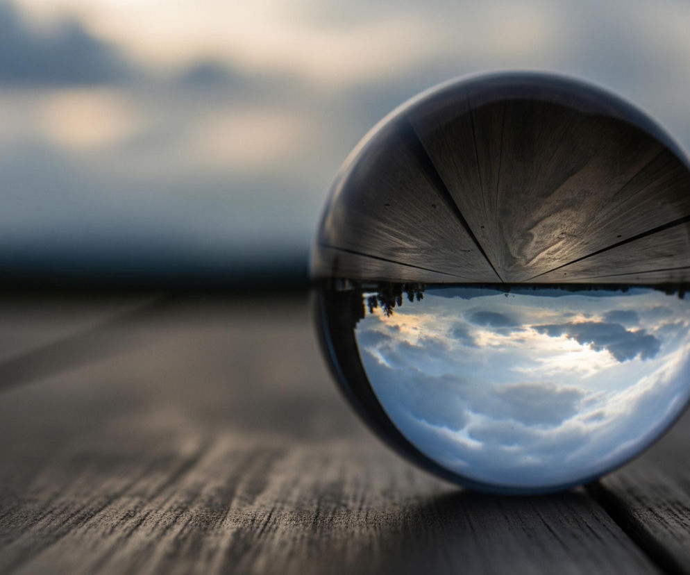 Szklana kula, ustawiona na ciemnym, drewnianym podłożu, dominuje środek kadru. Górna część kuli odbija drewnianą powierzchnię z widocznymi słojami i szczeliną, natomiast dolna część kuli ukazuje odwrócony obraz zachmurzonego nieba i odległego horyzontu. W tle, poza kulą, rozmyte niebo z szarymi i lekko oświetlonymi chmurami przechodzi w ciemny pas na horyzoncie.