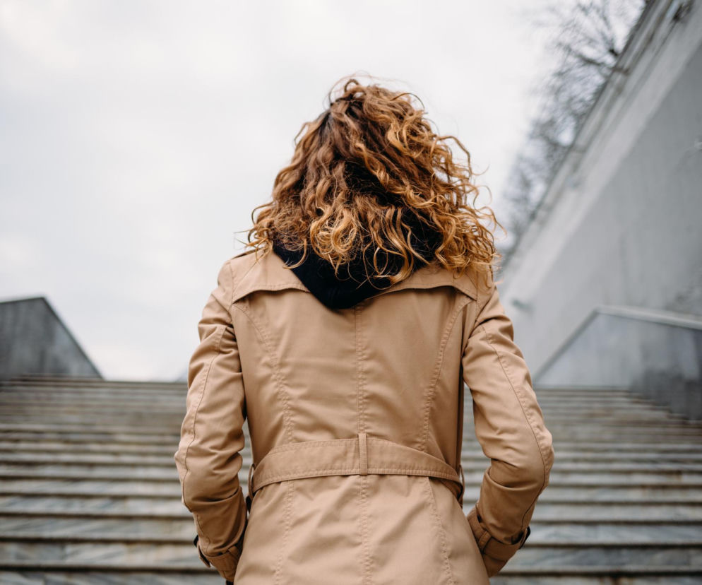 Kobieta o kręconych, rudych włosach w beżowym płaszczu, ze splecionymi rękami z tyłu, stoi na dole szerokich, kamiennych schodów, patrząc w górę. Obraz ilustruje tekst o zadyszce po wejściu na schody oraz kiedy to sygnał alarmowy, o czym przeczytasz na portalu Poradnik Zdrowie.