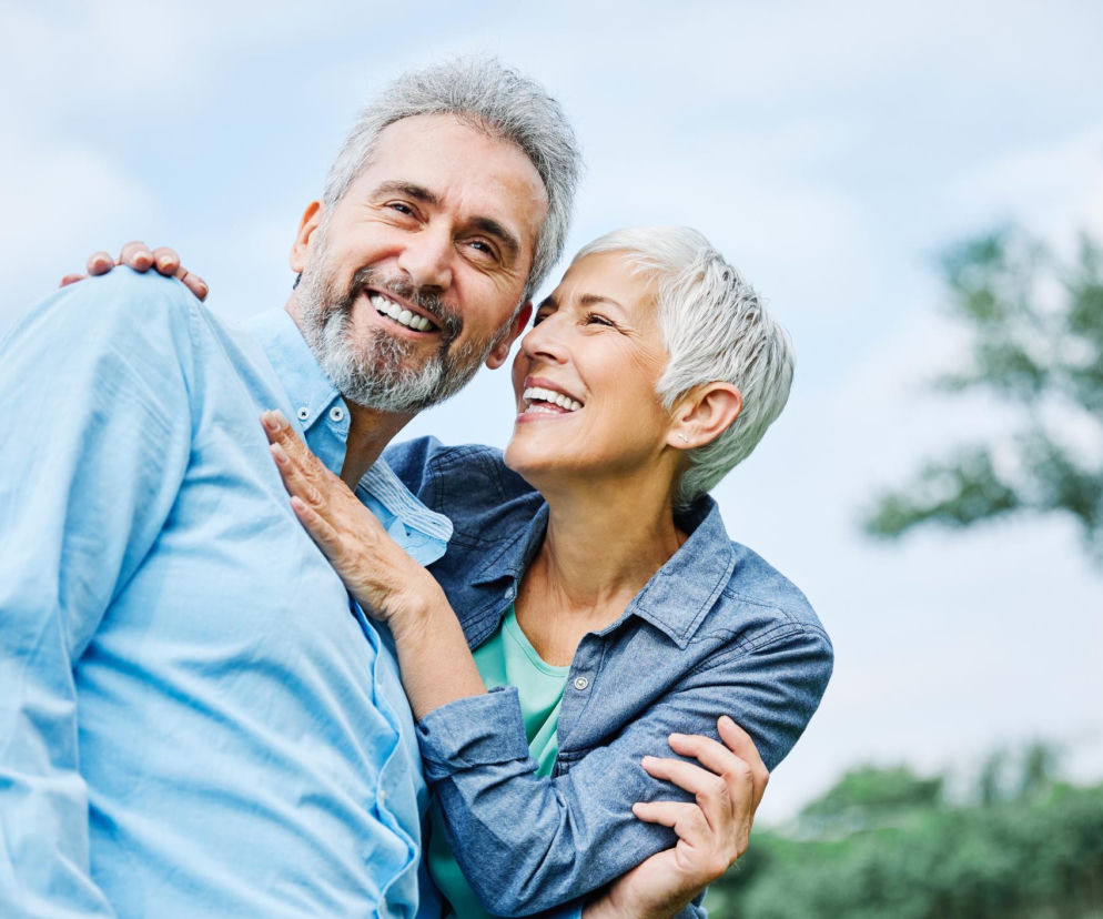Szczęśliwe, uśmiechnięte starsze małżeństwo obejmujące się na tle błękitnego nieba i zielonych drzew. Kobieta z siwymi włosami wpatruje się w mężczyznę z brodą. Więcej o zdrowiu po 40. na Poradnik Zdrowie.