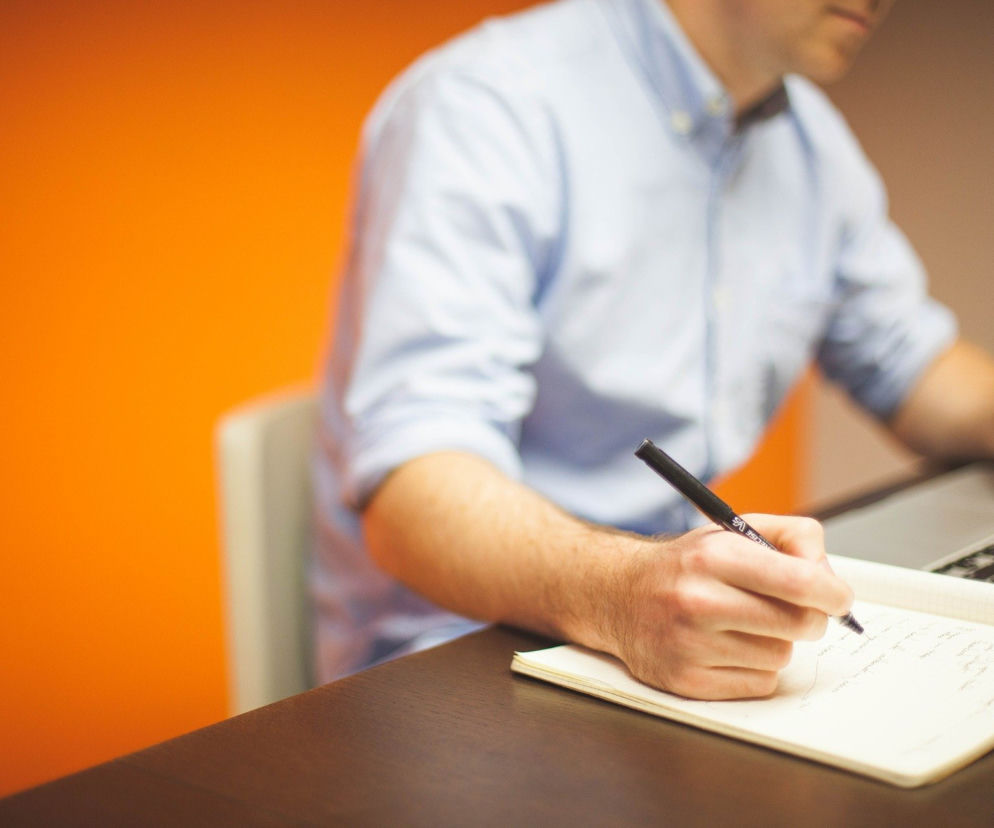 Mężczyzna w błękitnej koszuli piszący w notesie obok laptopa, symbolizujący pracę i formalizację umów od 2026 roku. Zdjęcie ilustruje zmiany na rynku pracy, o których możesz przeczytać na Super Biznes.