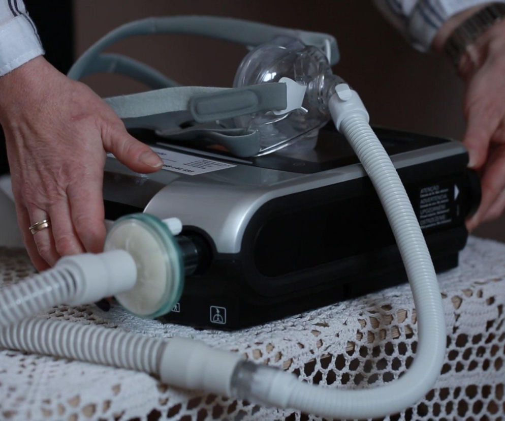 Urządzenie do domowej respiratoroterapii z maską tlenową i rurami, przygotowywane przez osobę. Na Poradniku Zdrowie dowiesz się więcej o problemach z dostępem do wentylacji mechanicznej.