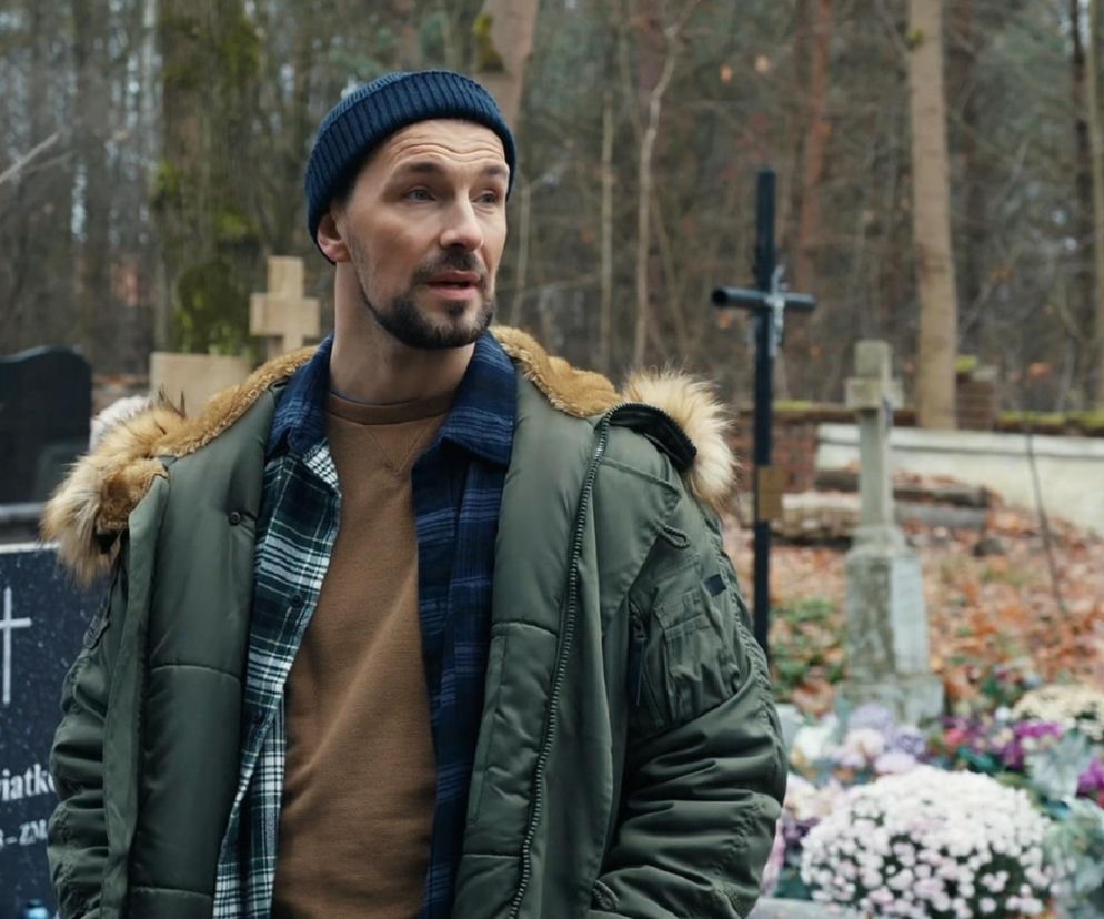 Marcin Kodur w zielonej kurtce z futrzanym kapturem i granatowej czapce stoi na cmentarzu, obok nagrobków. Jego wzrok jest skierowany w prawo, a wyraz twarzy sugeruje zamyślenie. To scena z serialu Barwy szczęścia, której opis znajdziesz na Super Seriale.