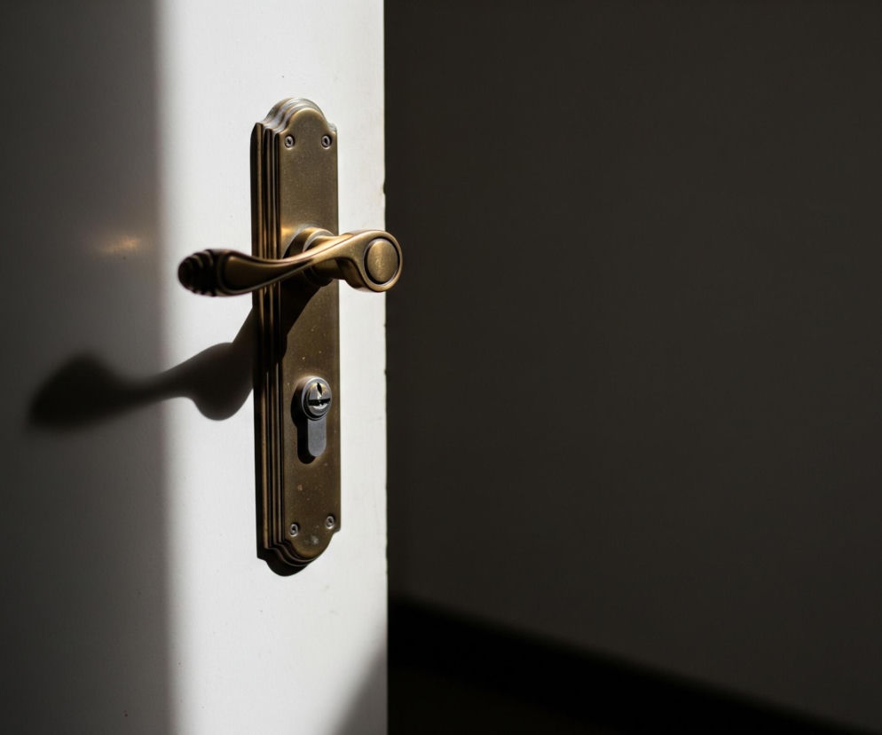 Antyczna, mosiężna klamka z ozdobną długą szyldą i wkładką zamka jest umieszczona na jasnych, białych drzwiach, lekko otwartych, ukazując ciemną przestrzeń. Światło padające z lewej strony tworzy wyraźny cień klamki oraz rzuca pionowy pas światła na drzwi. Prawa strona obrazu jest całkowicie zacieniona i ciemna.