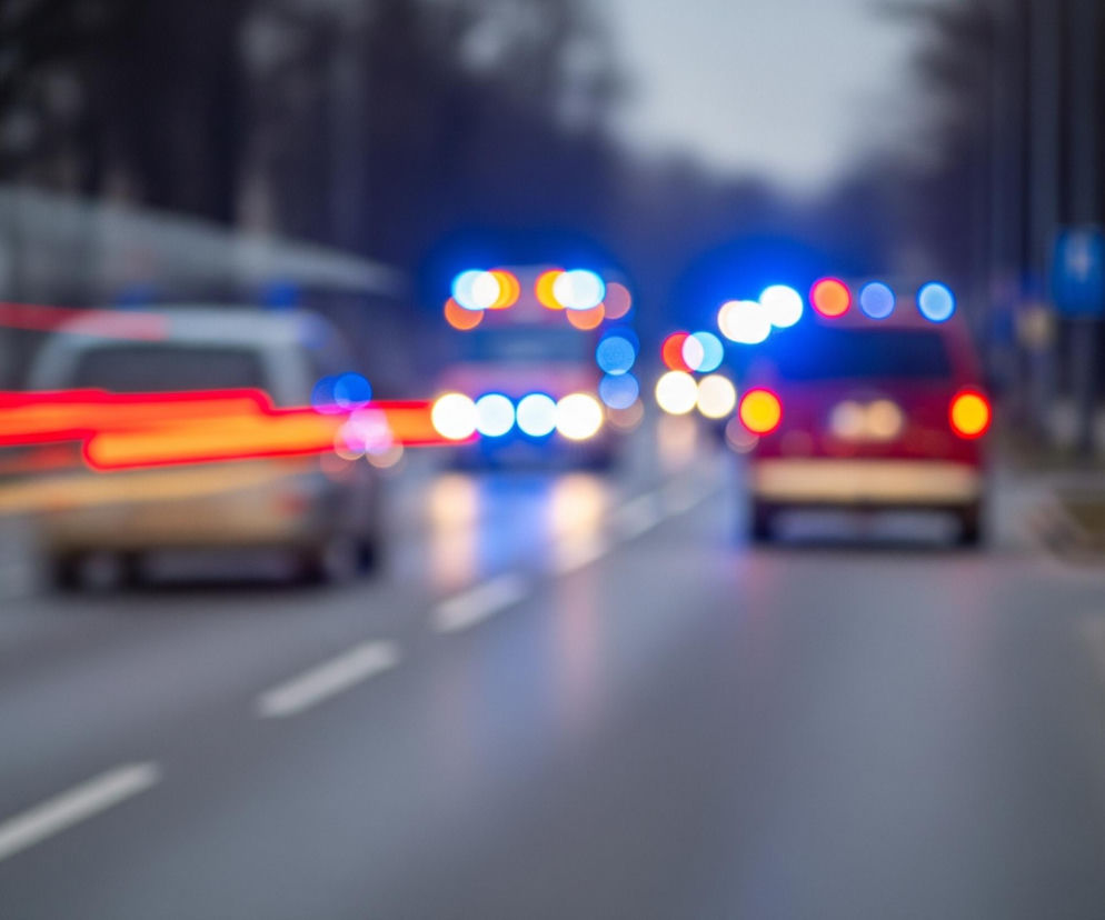 Główne obiekty na obrazie to trzy pojazdy uprzywilejowane jadące ulicą w kierunku od widza, z czego środkowy i prawy są czerwone, a lewy biały, wszystkie z włączonymi, rozmazanymi światłami awaryjnymi w kolorach niebieskim i czerwonym. Lewy pojazd pozostawia za sobą długie, poziome smugi czerwonego światła, sugerujące ruch i długie naświetlanie. Ulica jest szara z białymi przerywanymi liniami, a po prawej stronie widać chodnik i trawnik, natomiast tło jest rozmazane, ciemne i nieostre.