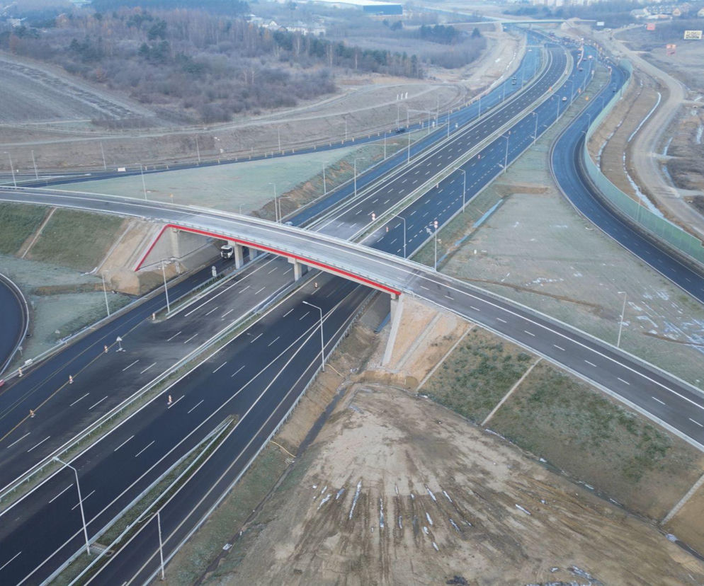 Widok z lotu ptaka na nowo otwartą Obwodnicę Metropolii Trójmiejskiej, z drogą ekspresową S7 biegnącą przez krajobraz. Widoczne są świeżo wytyczone jezdnie, węzły drogowe z wiaduktem o czerwonej balustradzie i liczne pasy ruchu, co jest kluczowe dla poprawy płynności ruchu w regionie, o czym przeczytasz na Architektura Murator Plus.