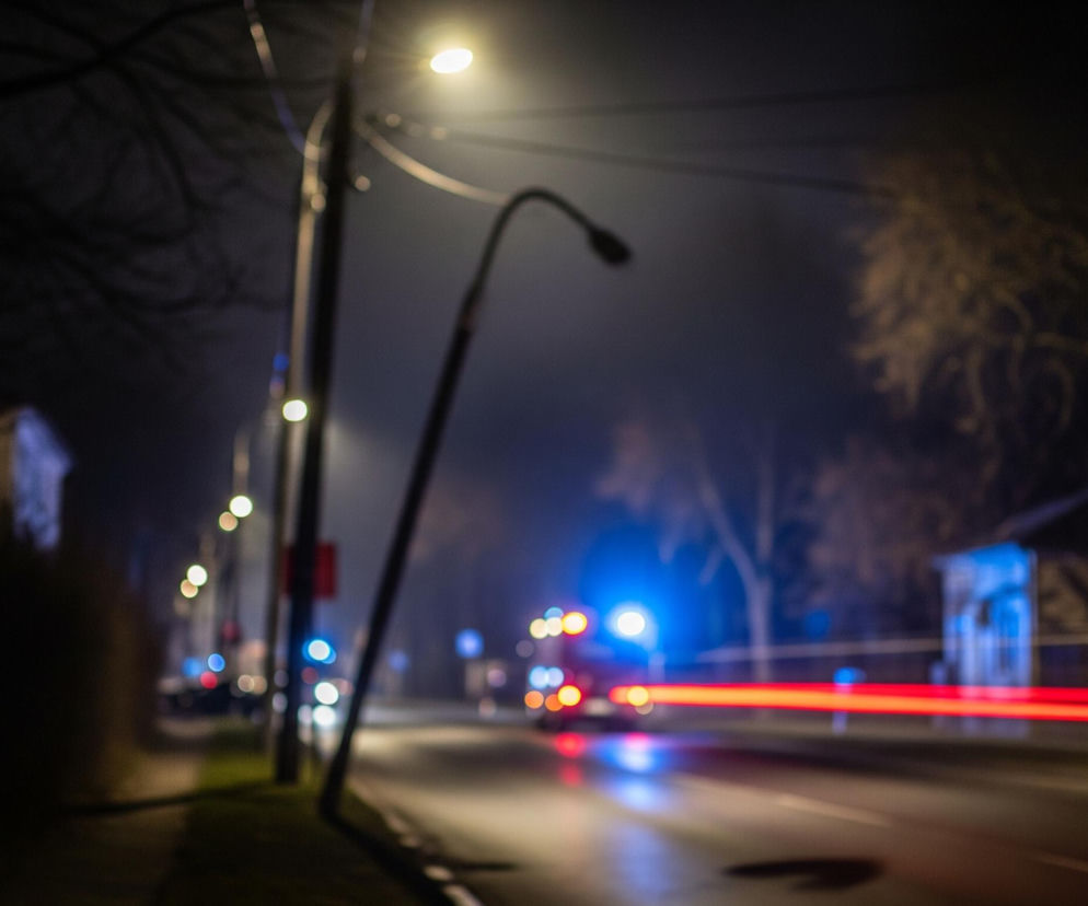 Oświetlona ulica nocą, sfotografowana z efektem rozmycia tła i światła. Na pierwszym planie po lewej stronie znajduje się słup, z którego wychodzą dwie linie energetyczne, a w górnej części widoczne jest jasnożółte światło lampy ulicznej. Po prawej stronie, nieco dalej, widać charakterystyczną, wygiętą lampę uliczną, której oprawa jest ciemna, a jej światło jest niewidoczne.

Ulica jest ciemna, mokra, ale w centralnej części widoczne są intensywne, czerwone, poziome smugi światła, które sugerują ruch pojazdu. Po prawej stronie, w tle, można dostrzec niebieskie światła, prawdopodobnie reflektory lub sygnały świetlne, oraz pomarańczowo-czerwone światła innych pojazdów. Po obu stronach ulicy znajdują się drzewa i niewyraźne zarysy budynków.
