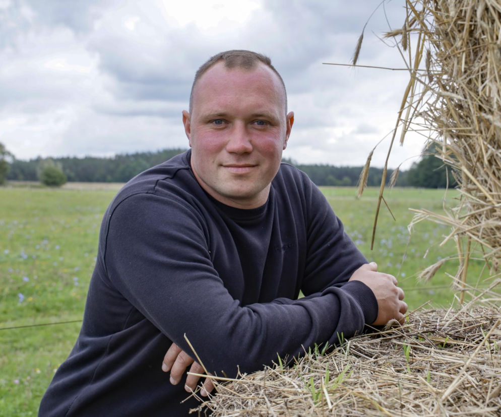 Uśmiechnięty rolnik Mariusz opiera się o snop siana na tle łąki. O jego obawach w programie przeczytasz na SE.
