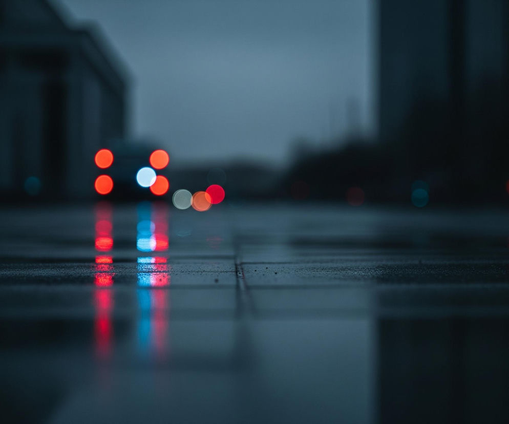 Na pierwszym planie, w dolnej części obrazu, widoczna jest mokra powierzchnia chodnika lub ulicy, złożona z ciemnych, prostokątnych płyt z widocznymi fugami. Na tej powierzchni odbijają się rozmyte światła w kolorach czerwonym, niebieskim i białym. Głębiej w kadrze, na rozmytej ciemnej drodze, widoczne są okrągłe światła samochodów: pięć czerwonych świateł pozycyjnych lub hamowania, jedno jasne białe światło oraz kilka mniejszych, mniej wyraźnych świateł w różnych odcieniach czerwieni i zielonkawego błękitu. Tło jest niewyraźne, zarysowują się na nim ciemne, geometryczne kształty budynków pod szarym, zachmurzonym niebem, co nadaje całości ponury i miejski charakter.