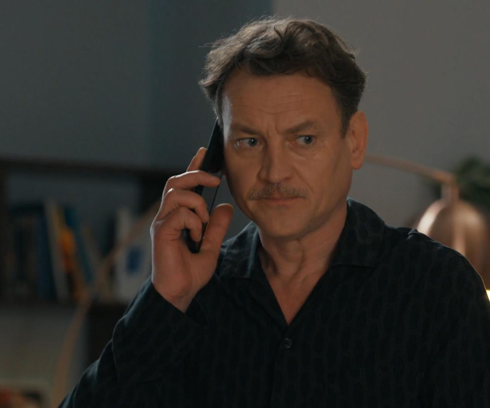 Paweł Ławrynowicz jako Stefaniak rozmawia przez telefon, patrząc w prawo, w tle zamglone wnętrze. Aktor ma rozczochrane włosy, wąsy i koszulę w ciemny wzór. Więcej o losach bohatera znajdziesz na Super Seriale.