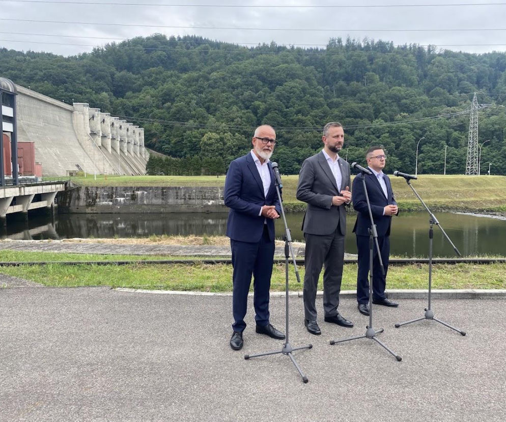 Trzej mężczyźni w garniturach przy mikrofonach na tle zapory wodnej. O inwestycji Tauronu pisze Eska Nowy-Sącz.
