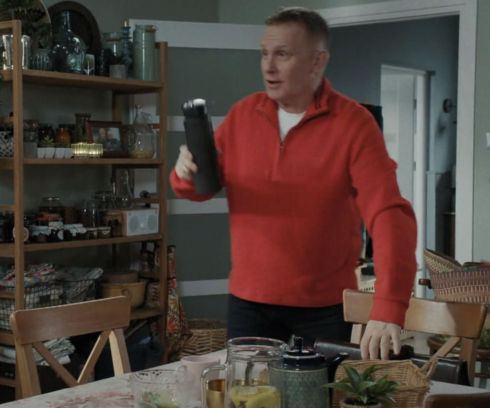 Robert Moskwa jako Artur Rogowski z M jak miłość w czerwonej bluzie z zamkiem, trzymający czarną butelkę z odżywką białkową. Zmiana stylu życia Artura dla Joanny to główny temat w Super Seriale. W tle drewniany regał z przetworami i innymi akcesoriami kuchennymi.