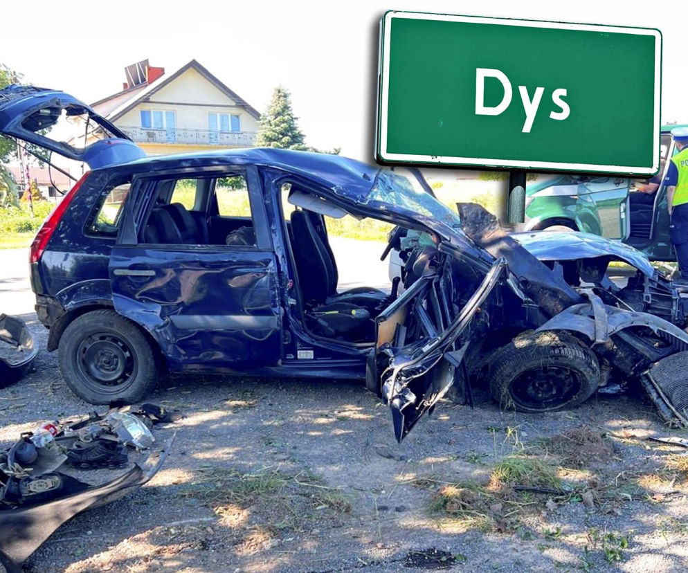 Zniszczony, granatowy Ford Fiesta po wypadku drogowym, z urwanym przodem i otwartymi drzwiami, leży na poboczu drogi w Dysie. Obok samochodu leżą elementy karoserii, w tle widać policjanta i tablicę z nazwą miejscowości. O tragicznym wypadku przeczytasz na naszym portalu.
