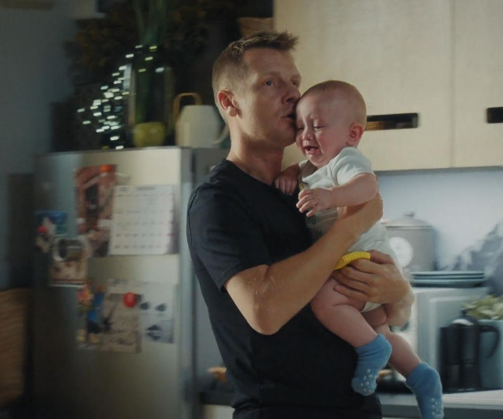 Paweł Zduński (Rafał Mroczek) w czarnej koszulce, przytula i próbuje pocieszyć płaczącego Antosia (Mark Myronenko) w kuchni, odcinek M jak miłość. Tę scenę rozpaczy po śmierci Franki zobaczycie na portalu Super Seriale.