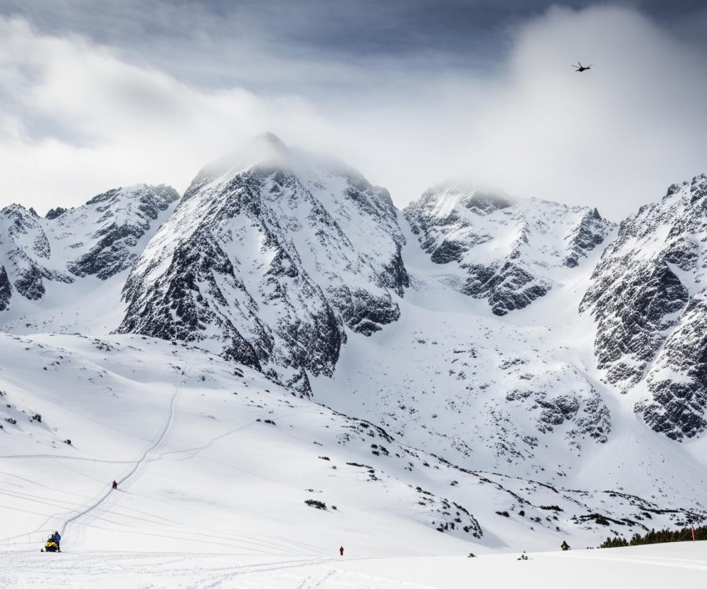 Szeroki plan przedstawia ośnieżone, skaliste góry, których szczyty giną w chmurach. Dominują zimne barwy: biel śniegu, szarość skał oraz niebo w odcieniach bieli i błękitu. Na pierwszym planie widać szeroką dolinę pokrytą gęstą warstwą śniegu, z kilkoma śladami nart lub desek śnieżnych i odległymi postaciami w jaskrawych, ciepłych kolorach. Jeden z obiektów to żółty skuter śnieżny z siedzącą na nim postacią, a w tle widać czarny helikopter lecący pod szarymi chmurami.