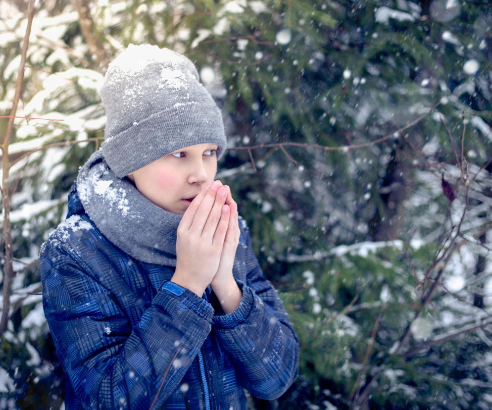 Dziecko w szarej czapce i szaliku, w zimowej, niebieskiej kurtce, ogrzewające dłonie ustami podczas mroźnego, śnieżnego dnia, ukazujące ryzyko odmrożeń. Więcej o ochronie przed odmrożeniami na Poradnik Zdrowie.