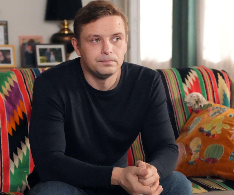 Marek Molak jako Hubert Pyrka, siedzący na kolorowej kanapie z charakterystycznym wzorem etnicznym, patrzący w dal z zatroskaną miną, z dłońmi splecionymi. W tle półka ze zdjęciami. Więcej o serialu Barwy szczęścia znajdziesz na portalu Super Seriale.