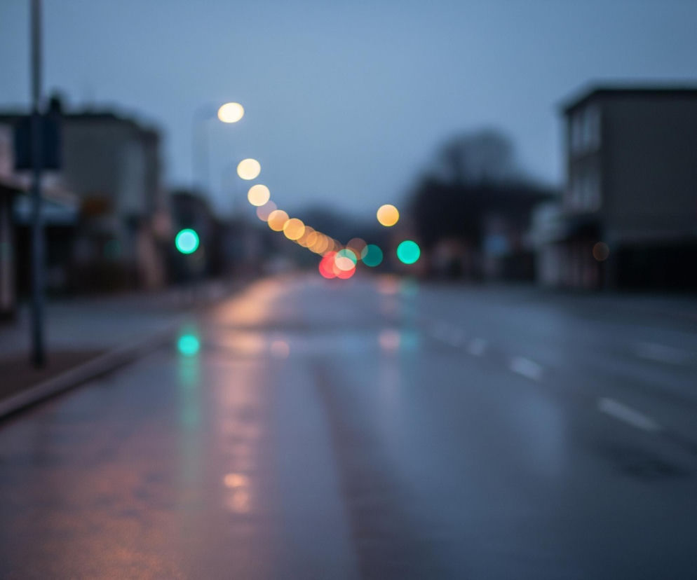 Uliczna perspektywa w miękkim, rozmazanym ujęciu przedstawia drogę z odblaskami świateł na mokrej nawierzchni. Po obu stronach widoczne są niewyraźne zarysy budynków, a w oddali widać linię słupów z żółtymi i czerwonymi punktami światła, tworzącymi efekt bokeh. Dominują barwy szarości i błękitu w tle, przełamane intensywną zielenią świateł sygnalizacji świetlnej i ciepłymi odcieniami lamp ulicznych.