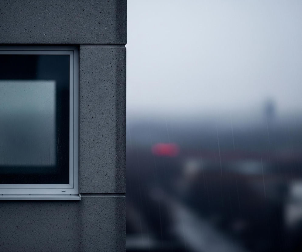 Lewą część kadru zajmuje fragment szarej, nakrapianej ściany budynku, w której osadzone jest pionowe, prostokątne okno. Rama okna jest jasnoszara, a na jego szybie widoczne są pojedyncze krople deszczu. Pod oknem znajduje się wąski, jasnoszary parapet. Prawa strona obrazu przedstawia rozmyty, szarobłękitny pejzaż miejski podczas deszczu, z niewyraźnymi sylwetkami budynków i drzew w tle oraz smugami opadów. Wśród rozmytych kształtów tła wyróżnia się jeden intensywnie czerwony punkt świetlny.