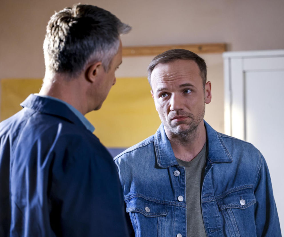 Dwóch mężczyzn, Łukasz Płoszajski jako Artur i Maciej Mikołajczyk jako Janek z serialu Pierwsza miłość, stoi w pomieszczeniu. Janek, ubrany w jeansową kurtkę, patrzy z zamyśloną miną. Artur stoi tyłem, ubrany w niebieską koszulę. Więcej o ich losach w Super Seriale.